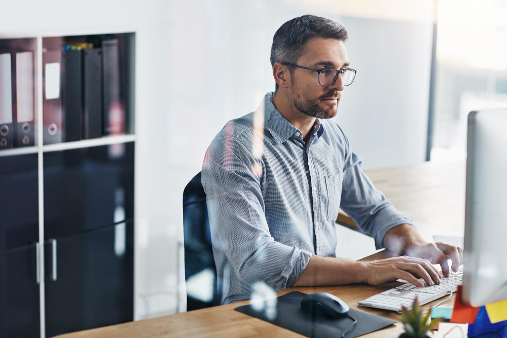 Homem de óculos focado a digitar no computador num escritório, a representar um especialista em intermediação de crédito a analisar o perfil de um cliente