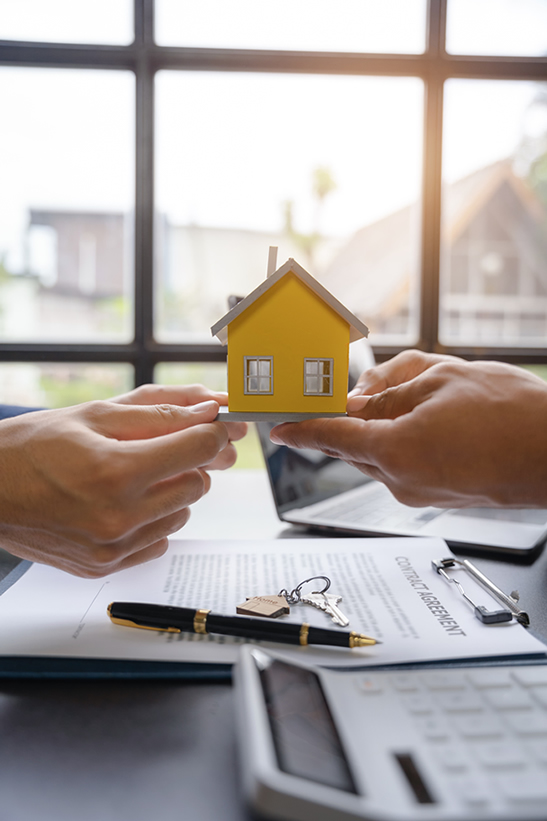 Duas pessoas a segurar uma casa em miniatura amarela sobre uma secretária com um contrato, chaves e uma calculadora, a representar a transferência de crédito habitação.